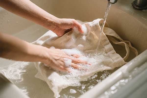 A person hand-washing a canvas tote bag in a sink with mild soap.