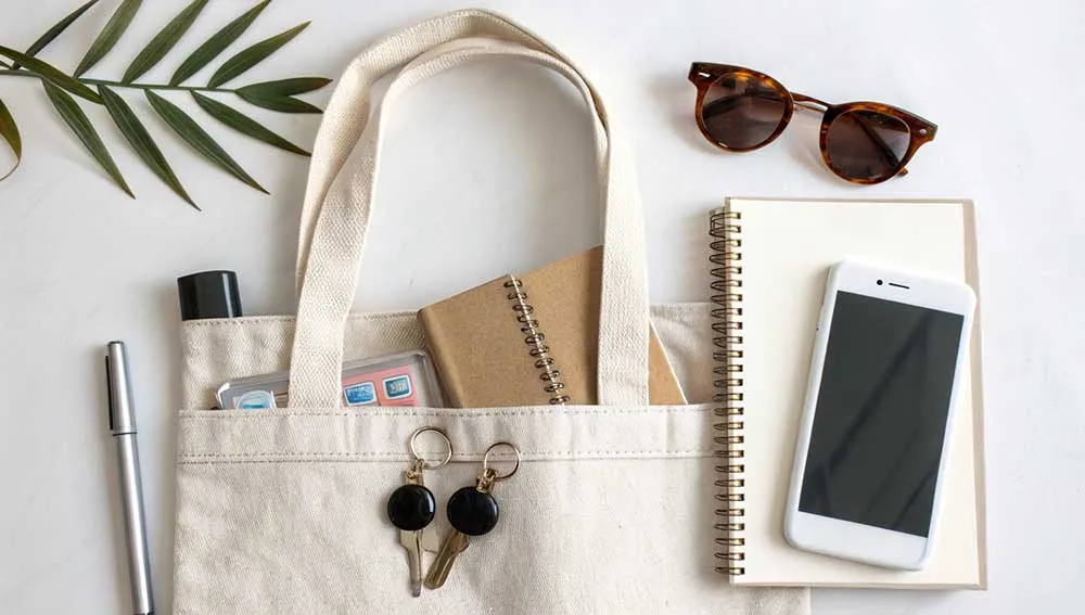 A neatly arranged flat lay of a tote bag containing a notebook, sunglasses, a smartphone, keys, a pen, and other daily essentials.