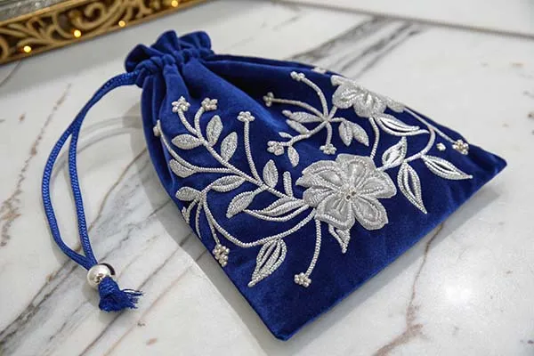 A royal blue velvet potli bag with silver floral embroidery and a drawstring closure, placed on a marble surface.
