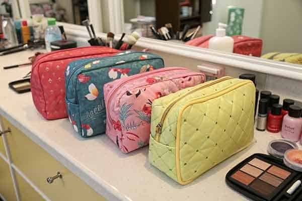 Four colorful quilted puffy makeup bags (red, blue with patterns, pink with patterns, yellow) lined up on a vanity counter, with various cosmetics around