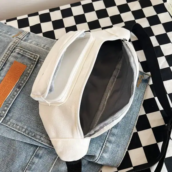 A casual white canvas shoulder bag with a black strap, open to show its interior, placed on a checkered cloth with a pair of jeans underneath.