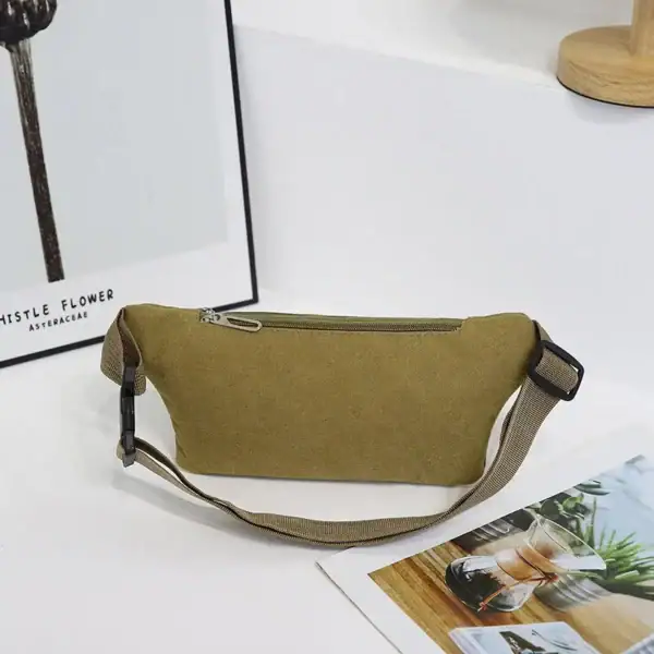 A casual canvas belt bag in olive color with a zipper and adjustable strap placed on a white surface with a framed artwork and a magazine nearby.