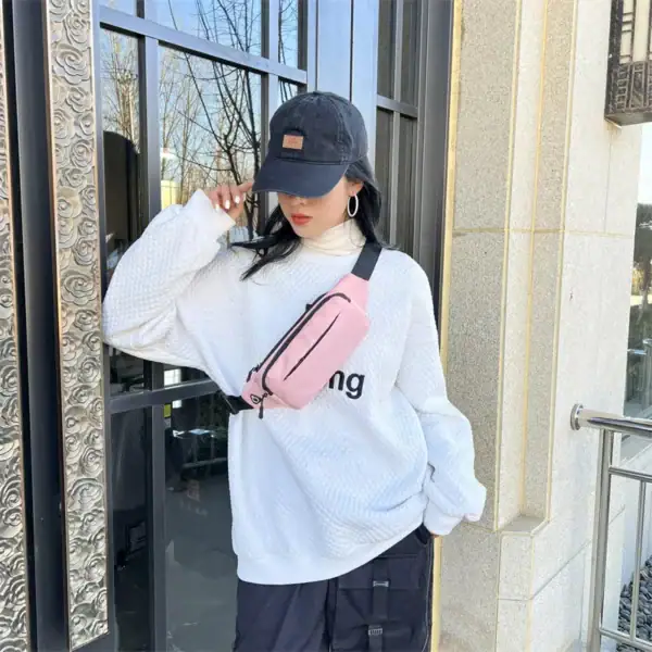 A person wearing a white textured sweater, black cap, and a pink sling bag while posing near a decorative outdoor glass door with tree reflections.