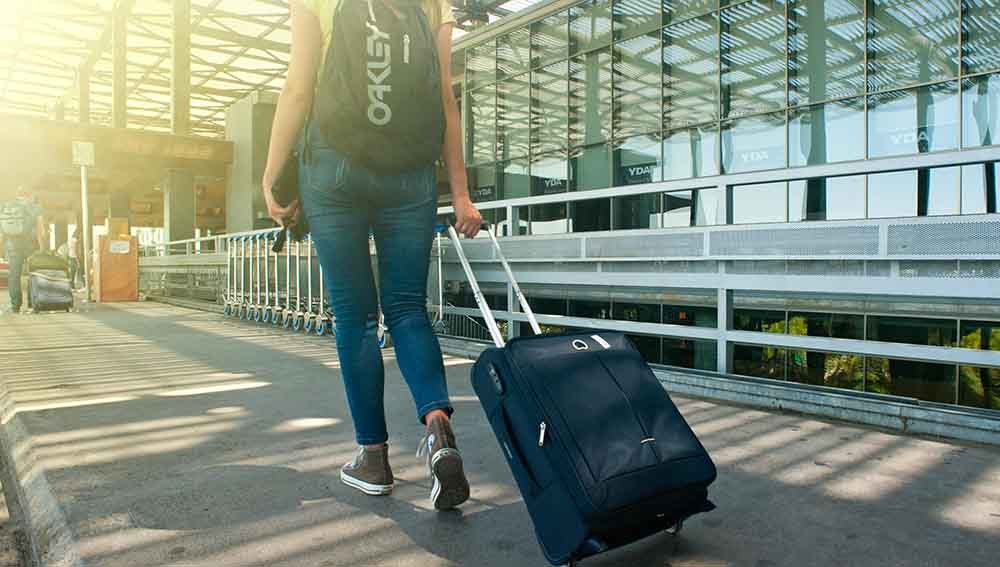 A woman walking with a suitcase at the airport with an oxford backpack and a sunny background.