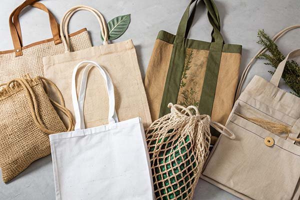 Assorted eco bags in different materials (jute, canvas, etc.) laid out on a light surface, showcasing diverse styles for eco-friendly use.