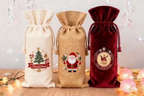 Three printed wine bags in different materials (canvas, jute, velvet) with Christmas designs, displayed on a wooden surface with festive lights and decorations, perfect for holiday gifting.