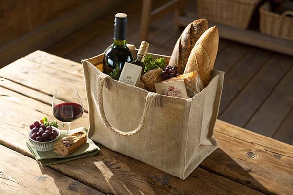 Sturdy jute bag with reinforced handles, filled with a bottle of wine and small food items.