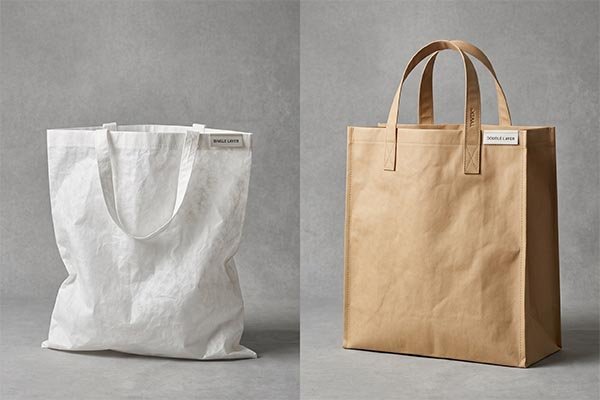 Side-by-side construction comparison showing a soft, white single-layer Tyvek tote bag on the left and a rigid, brown double-layer Tyvek tote bag on the right.
