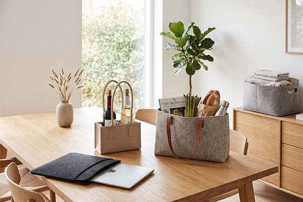 A felt wine carrier holding two bottles next to a felt grocery bag and a felt laptop sleeve on a wooden dining table.