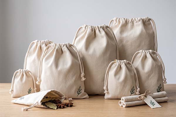 Custom canvas drawstring bags in various sizes with a green leaf logo, including rolled bags tied with custom tags, arranged on a table.