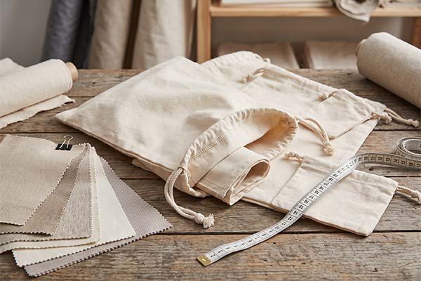 Unfinished eco-friendly cotton canvas dust bags on a wooden workbench, arranged with a measuring tape and a selection of beige and grey fabric swatches for material and dimension reference.