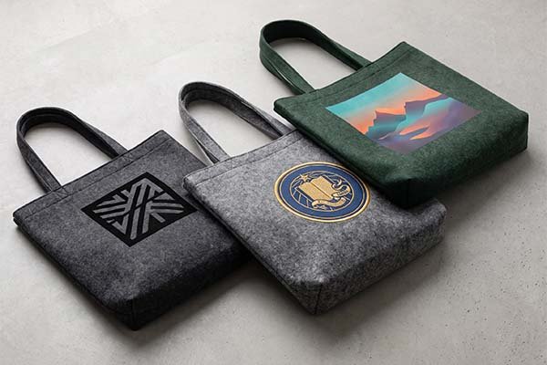 Three felt bags in charcoal, light grey, and green illustrating different logo formats and design limits on fuzzy material, featuring solid black prints, embroidered patches, and colorful printed blocks.