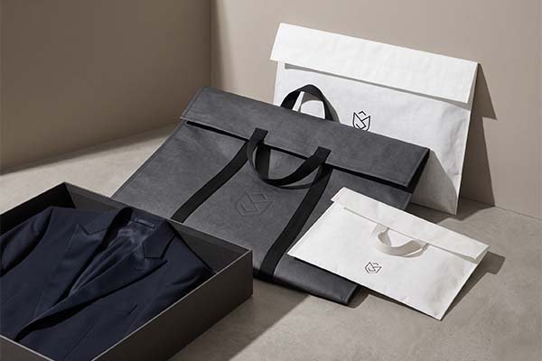 Tyvek garment packaging system featuring a large dark gray tote bag and white organizer bags, placed next to a navy blue custom suit in a premium paper box.