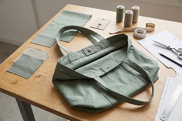 Mint green canvas tote bag sample on a workbench with matching fabric swatches, thread spools, and paper patterns.