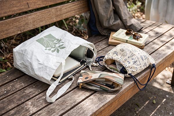 Reusable Tyvek merchandise styled for everyday use, featuring a tote bag, drawstring pouch, and zipper case resting on a wooden park bench with personal items.